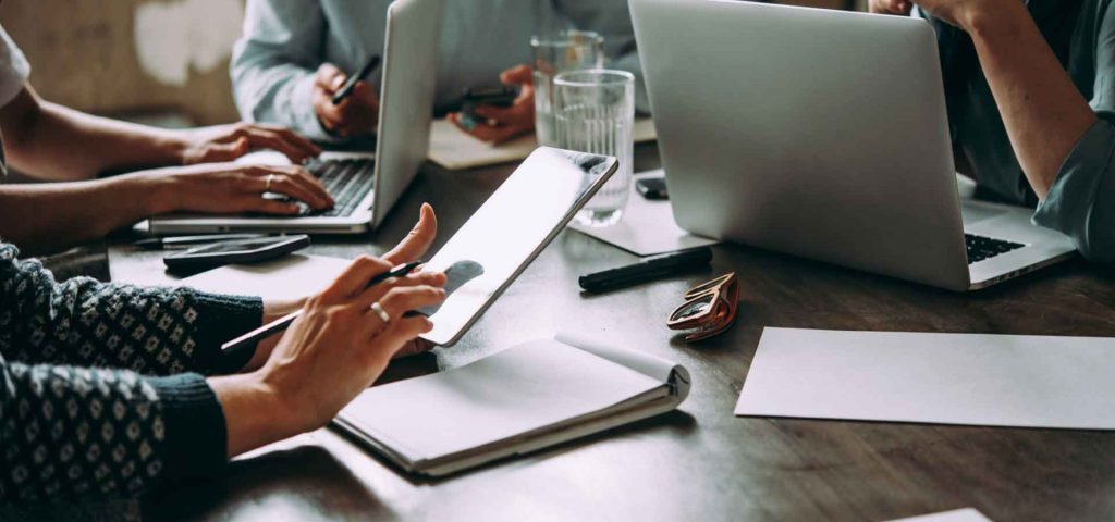 Eine Gruppe von Mitarbeitern sitzt bei einem Meeting an dem Tisch. Eine Frau tippt etwas auf ihrem Tablet ein.